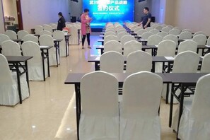A conference room with rows of tables and chairs, a stage with a blue screen, and a person standing on the stage. - Huangsheng Hotel (Wuchuan)