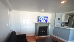 A living room with a black sofa, a coffee table, a TV on a stand, a fireplace, and a kitchen area with a sink and cabinets. - Direct ocean view 2 bed 2 bath 36 ft deck walk to beach. (Hull)