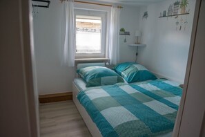 Un dormitorio pequeño con una cama, un ventanal con cortinas, una mesita de noche con lámpara y un estante en la pared con plantas.