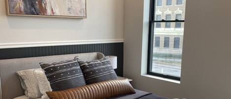 A modern bedroom with a large bed, a headboard, a window with a view of buildings, and a painting on the wall.