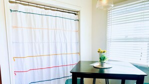 A dining area with a table, two chairs, a curtain with colorful stripes, a potted plant, and a window with blinds. - Midtown Ventura Family & Pet Friendly Cottage (Ventura)