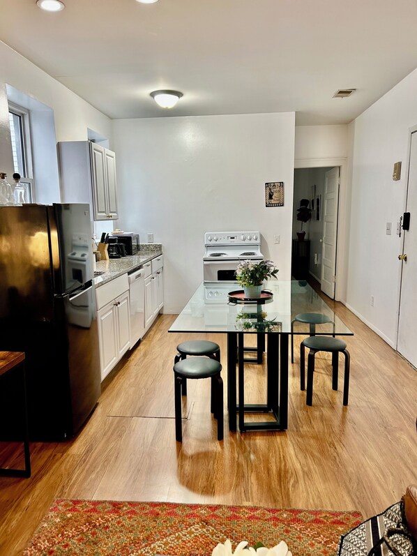 A modern kitchen with white cabinets, a black refrigerator, a glass dining table with four stools, and a wooden floor.