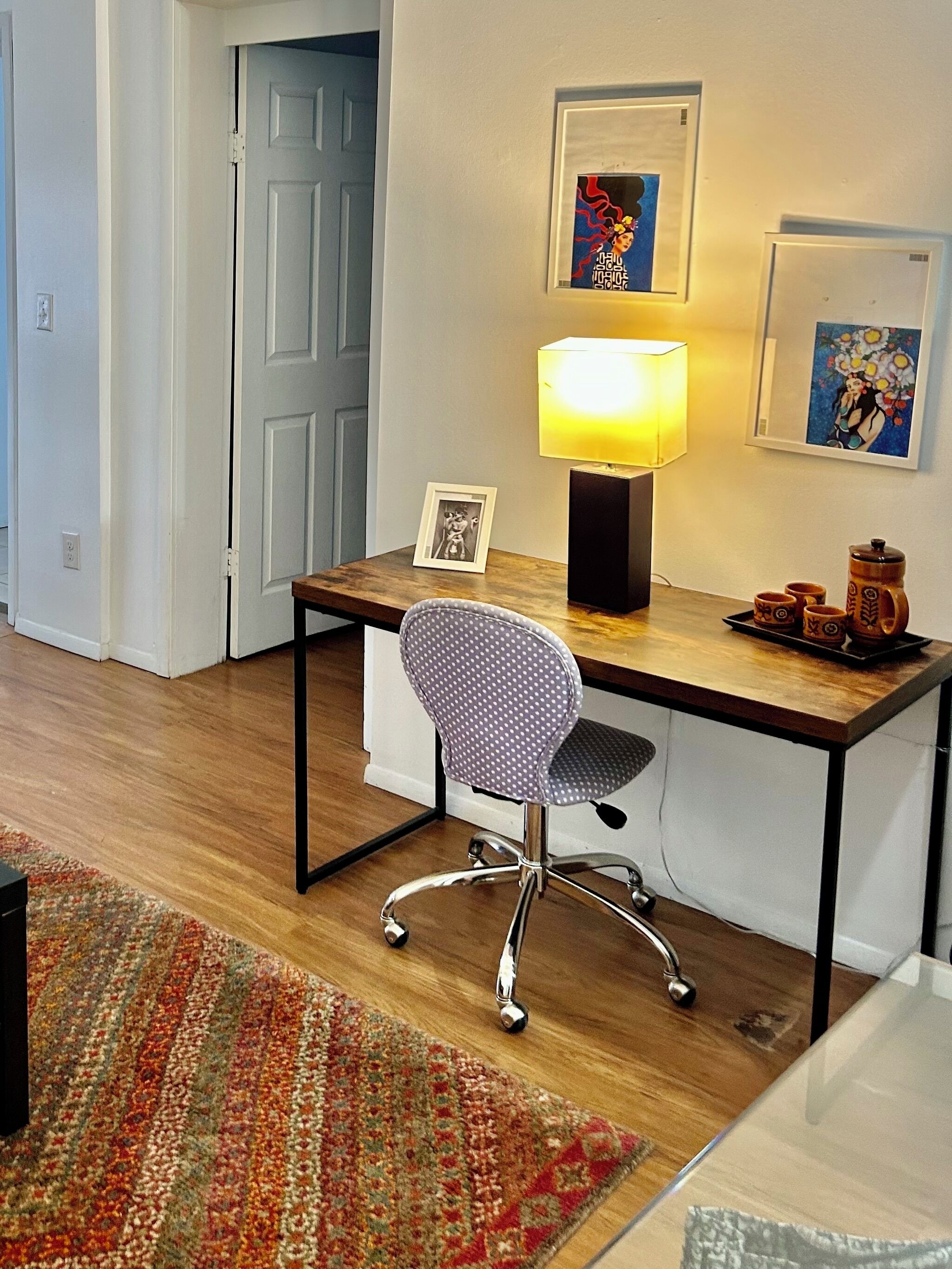 A room with a wooden desk, a chair, a lamp, a framed picture, and a small table with cups and a teapot.