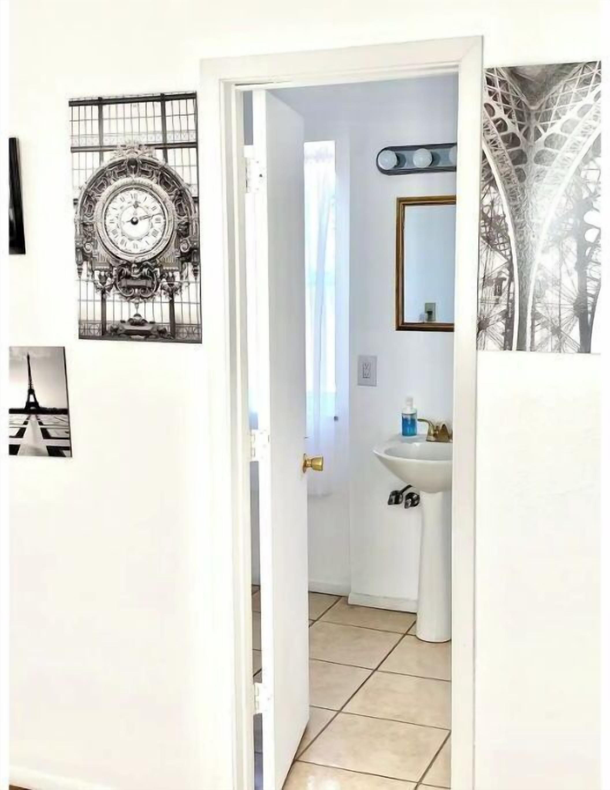 Un couloir étroit avec une porte blanche donnant sur une salle de bain. La salle de bain est équipée d’un lavabo blanc et d’un miroir au-dessus. Sur le mur, on trouve une grande horloge décorative et un tableau encadré représentant la Tour Eiffel.