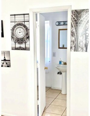 A narrow hallway with a white door leading to a bathroom. The bathroom has a white sink and a mirror above it. On the wall, there is a large decorative clock and a framed picture of the Eiffel Tower.