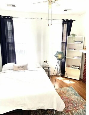A bedroom with a bed, a bedside table, a lamp, a bookshelf, and a window with curtains.