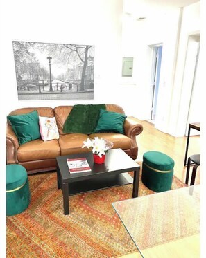 A living room with a brown leather sofa, green cushions, a black coffee table with a book and flowers, and a patterned rug.