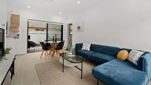 A modern living room with a blue sofa, a glass coffee table, and a dining area with a black table and chairs. - Stroll to beach, cafes & restaurants (BOOKER BAY)