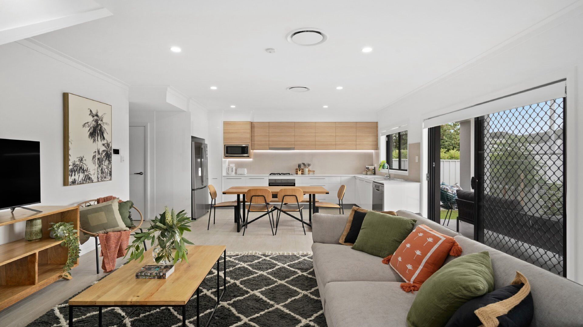 A modern living room with a sofa, coffee table, and TV. There is a kitchen area with wooden cabinets and a dining table. The room has large windows and a view of the outdoors.