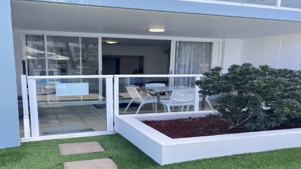 Een modern gebouw met een glazen deuropening die uitkomt op een balkon met witte stoelen en een tafel.