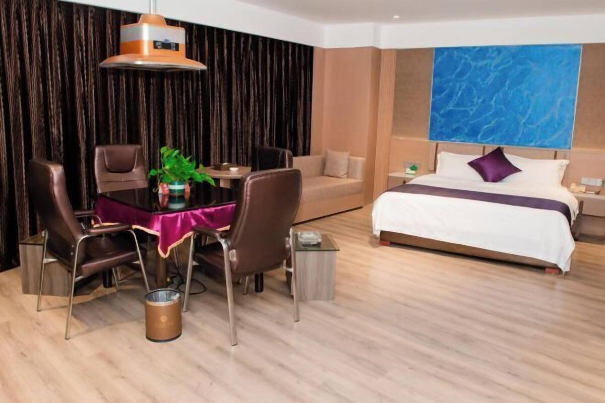 A modern hotel room with a bed, a dining table with chairs, a sofa, and a large blue artwork on the wall.