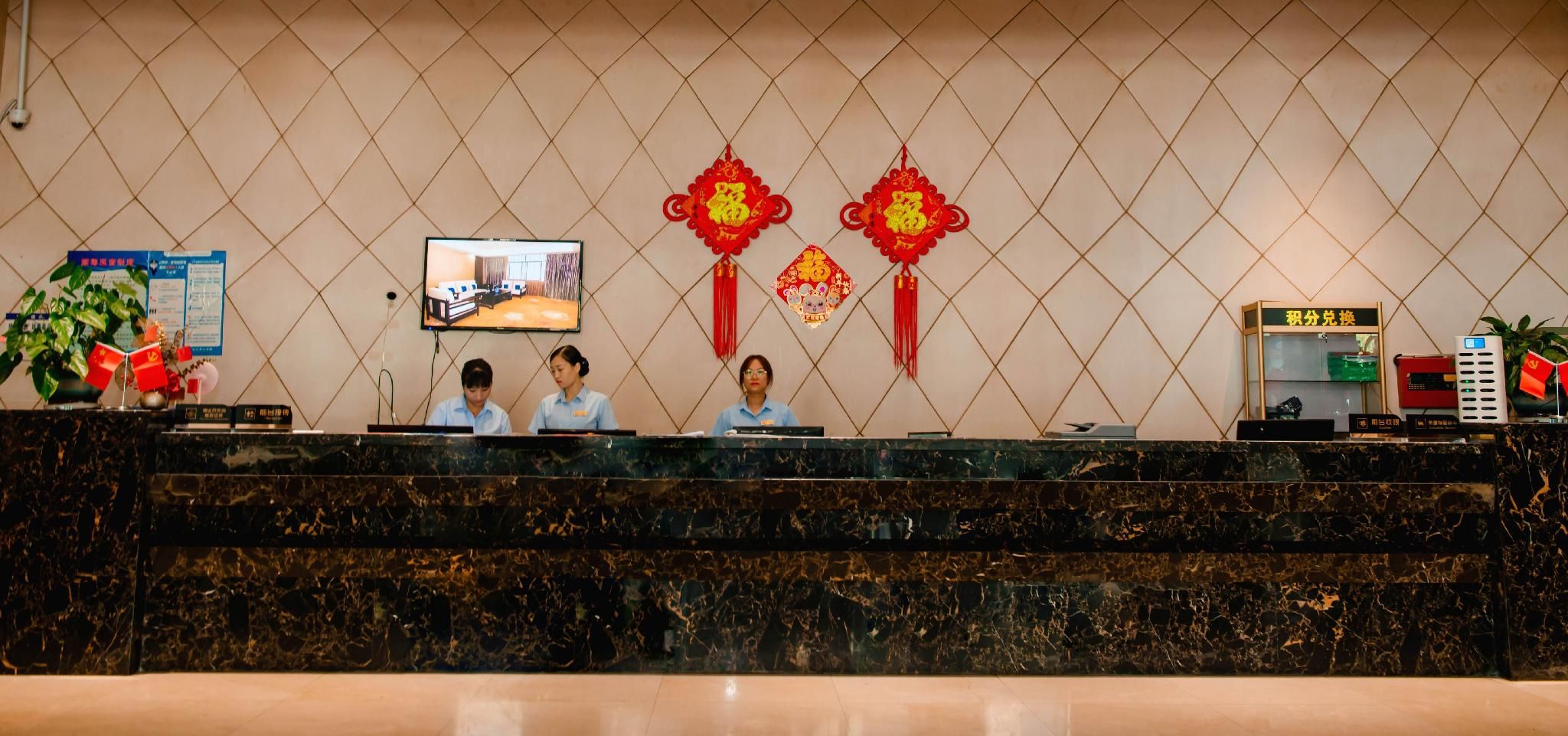 A hotel reception desk with a marble counter, two receptionists, a television screen, and red decorative elements on the wall.