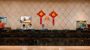 A hotel reception desk with a marble counter, two receptionists, a television screen, and red decorative elements on the wall.