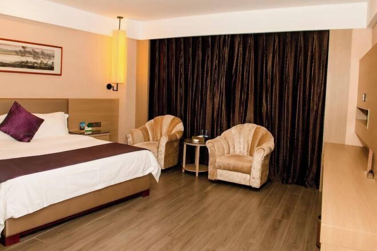 A hotel room with a large bed, two armchairs, a small table, and a dark curtain backdrop.