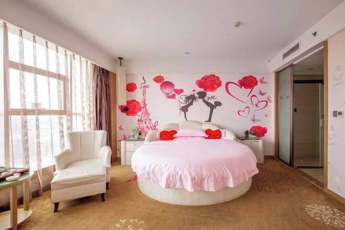 A bedroom with a round bed, a white armchair, and a large window with sheer curtains.