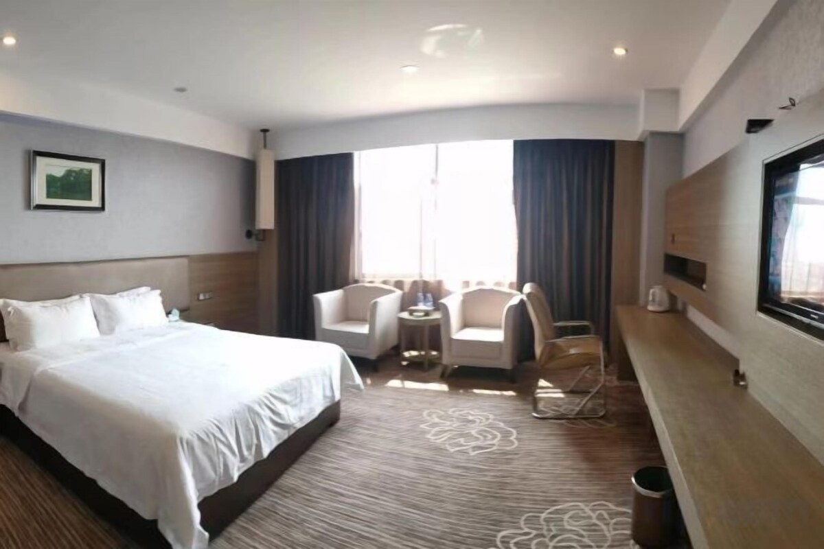 A hotel room with a large bed, a seating area with chairs, a small table, a TV mounted on the wall, and a window with curtains.