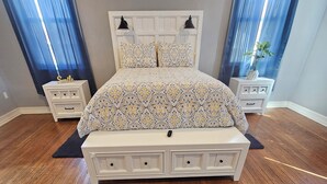 Un dormitorio con suelo de madera, una cama con colcha estampada, dos mesitas de noche y una mesita de noche con lámpara.
