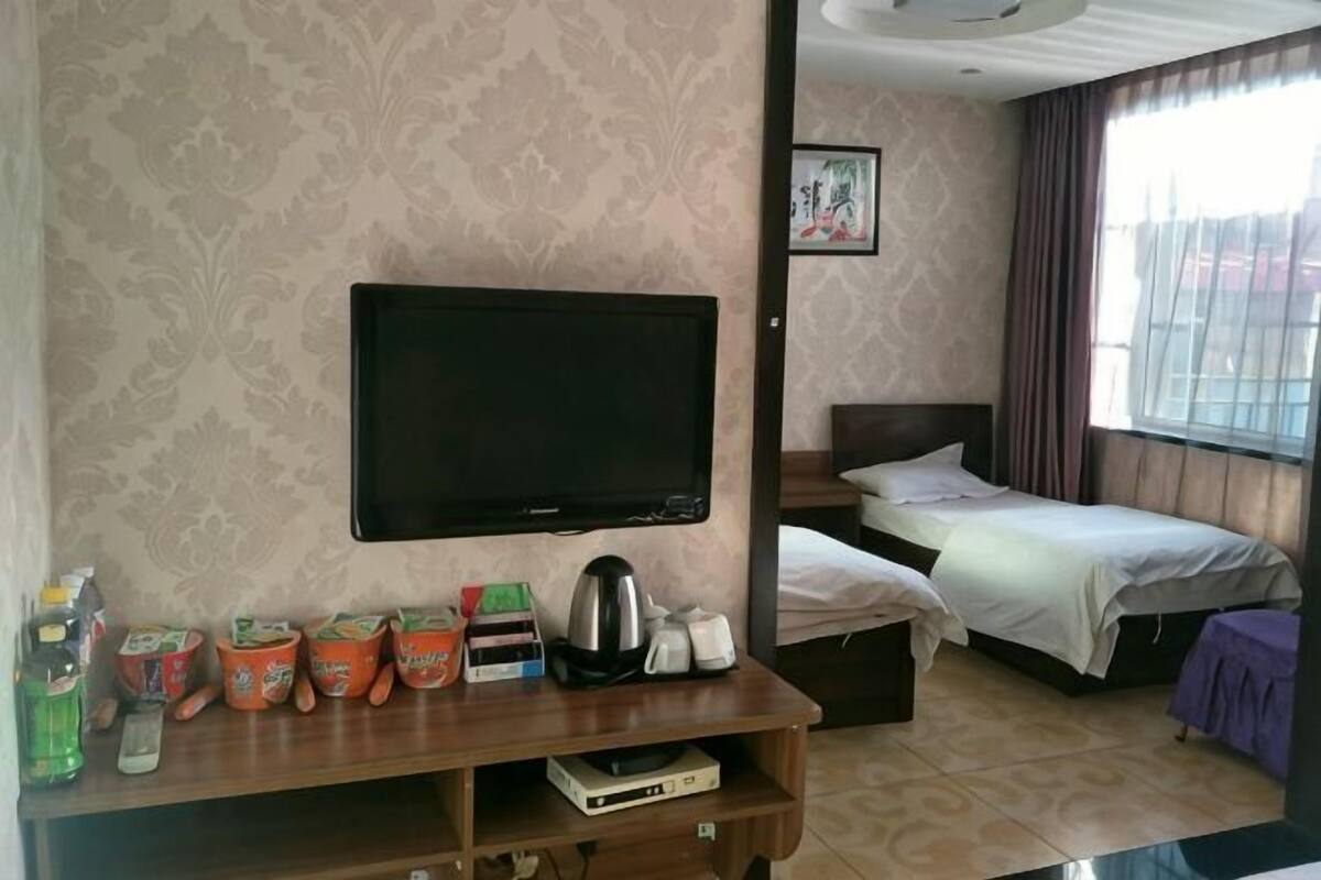 A hotel room with a flat-screen TV, two beds, a wooden TV stand with various items, and a window with curtains.
