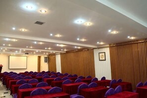 A conference room with rows of tables and chairs, a projector screen, and a wooden panel wall.