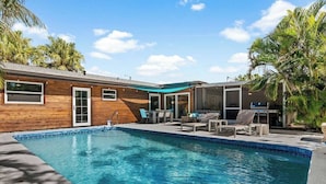 Un área de piscina moderna con revestimiento de madera, piscina azul cristalina y mobiliario exterior.