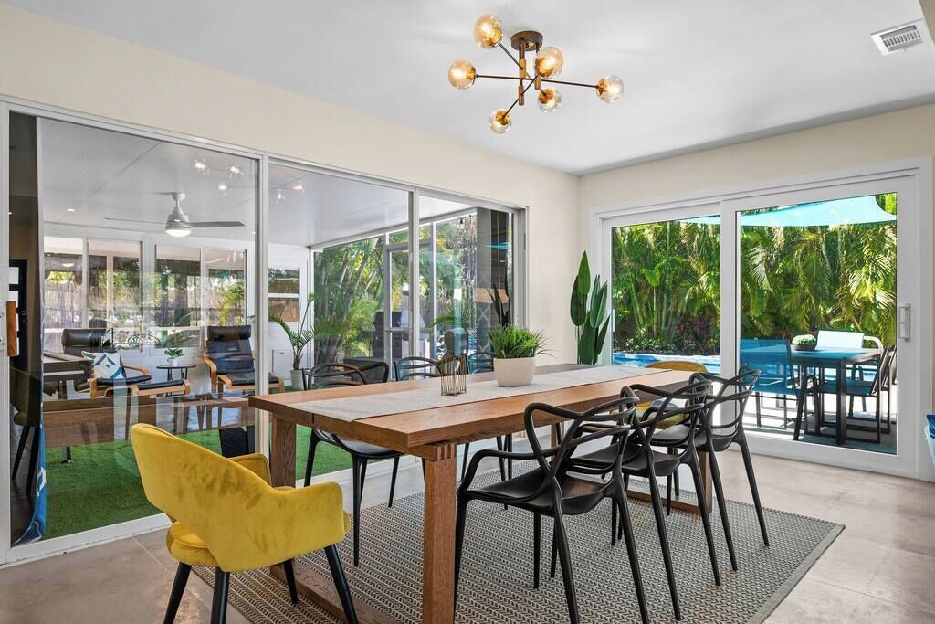 An open-plan living area with a wooden dining table, black chairs, and a yellow chair. There is a ceiling fan, a chandelier, and a view of a swimming pool and greenery outside.
