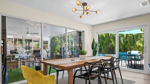 Un espacio habitable de concepto abierto con una mesa de comedor de madera, sillas negras y una silla amarilla. Hay un ventilador de techo, una araña y vistas a una piscina y vegetación al exterior.