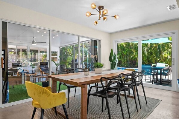 An open-plan living area with a wooden dining table, black chairs, and a yellow chair. There is a ceiling fan, a chandelier, and a view of a swimming pool and greenery outside. - Upscale. Heated Pool. Minutes to the Beach ! (Sarasota)