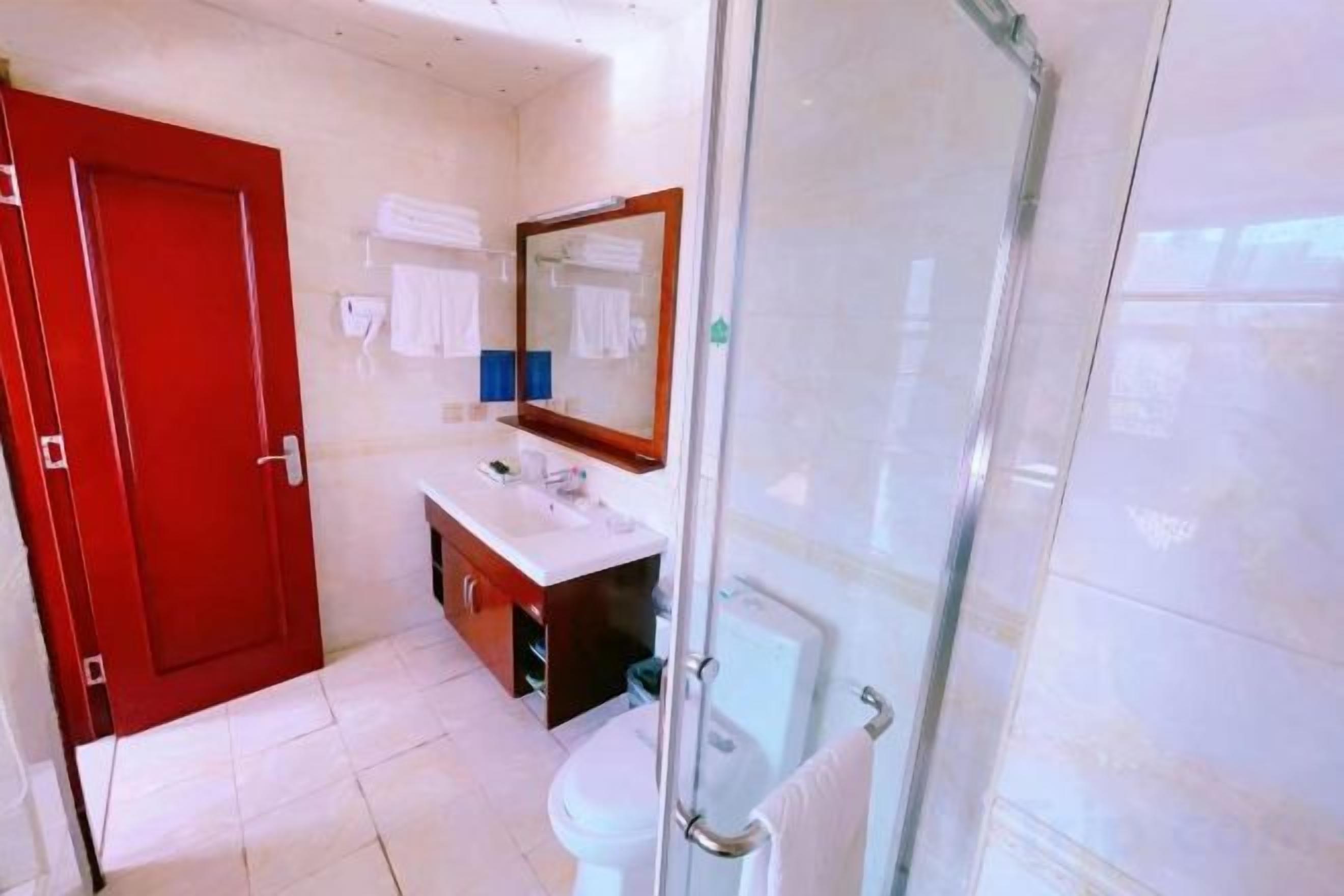Un baño con puerta roja, mampara de ducha de vidrio, lavamanos blanco y espejo.
