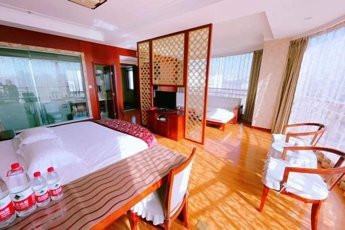 A hotel room with a bed, a TV, a wooden floor, and a view through large windows.