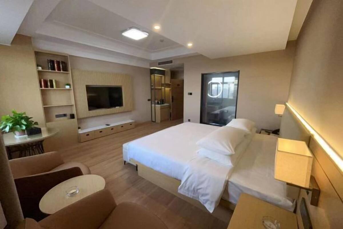 A modern hotel room with a large bed, a TV, a desk, a chair, and a bookshelf.