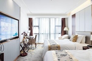 Habitación de hotel con dos camas, un escritorio con silla, televisor y ventanas amplias con cortinas.