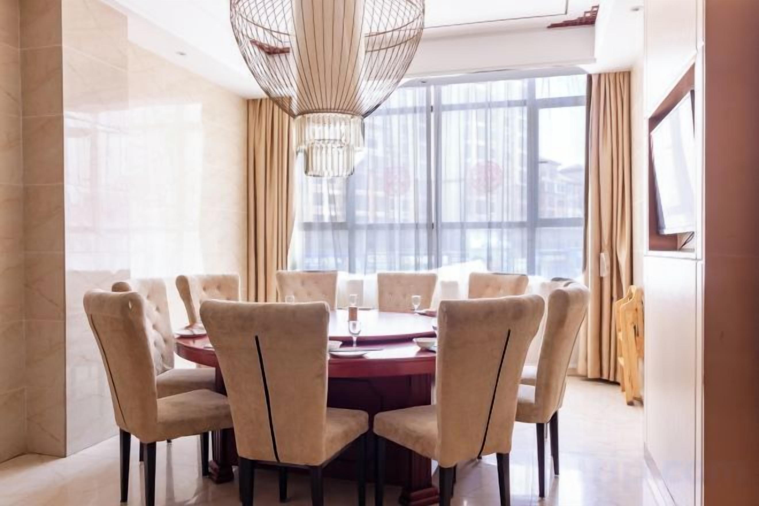A dining area with a round table and chairs, a chandelier, and a large window with curtains.