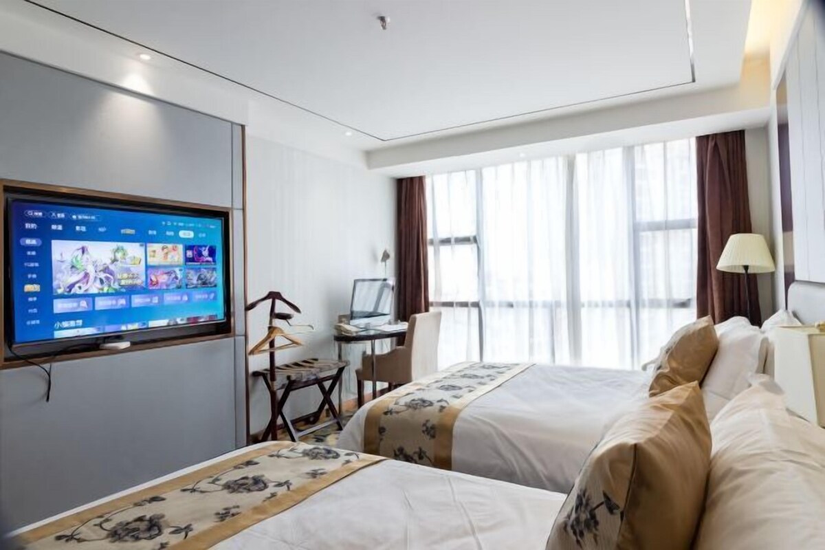 A hotel room with two beds, a desk with a computer, a TV mounted on the wall, and a large window with curtains.