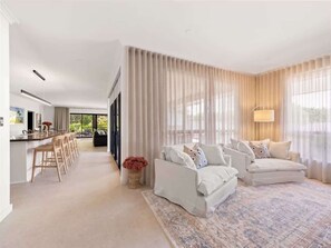 A modern living room with a sofa set, a dining area with bar stools, and a large window with sheer curtains.