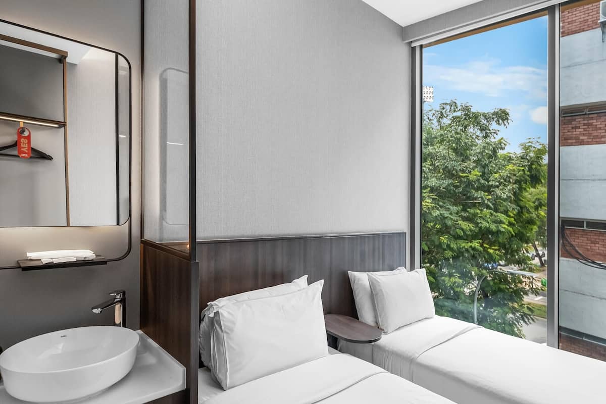 A modern hotel room with a large window, a built-in sink, a mirror, and a bed with white bedding.