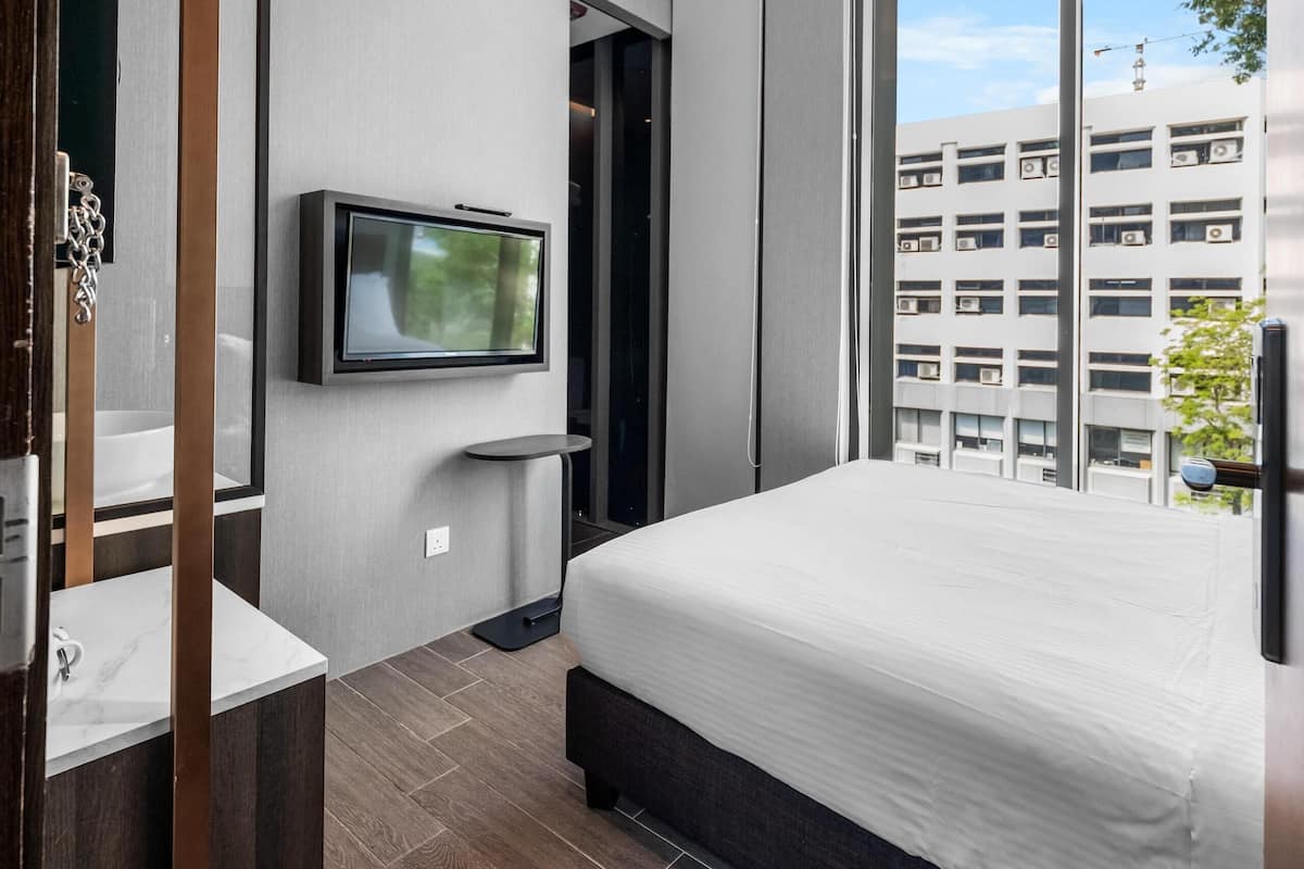 A modern hotel room with a large bed, a TV mounted on the wall, a bathroom with a mirror and sink, and a view of the city through large windows.