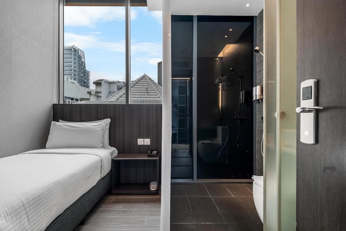 A modern hotel room with a bed, a small bedside table, a glass shower enclosure, and a view of the cityscape.