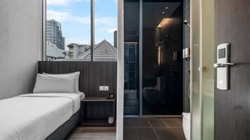 Quarto de hotel moderno com cama, mesa de cabeceira pequena, box de vidro para o chuveiro e vista para a cidade.