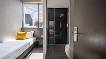 A modern hotel room with a bed, a small table, a window with a city view, a shower area, and a toilet.