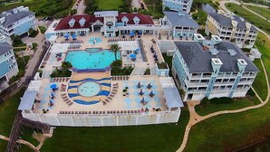 Eine Vogelperspektive eines Resorts mit mehreren Gebäuden, einem zentralen Poolbereich inklusive Liegestühlen und einer umlaufenden Rasenfläche.