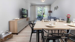 A modern dining area with a wooden table, black chairs, a TV on a wooden cabinet, and a view of the outdoors through large windows. - Bright Apartment with Balcony | Workspace (Kolobrzeg)