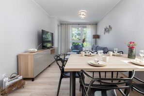 Ett modernt matområde med ett träbord, svarta stolar, en tv placerad på ett träskåp och utsikt över utomhusmiljön genom stora fönster.
