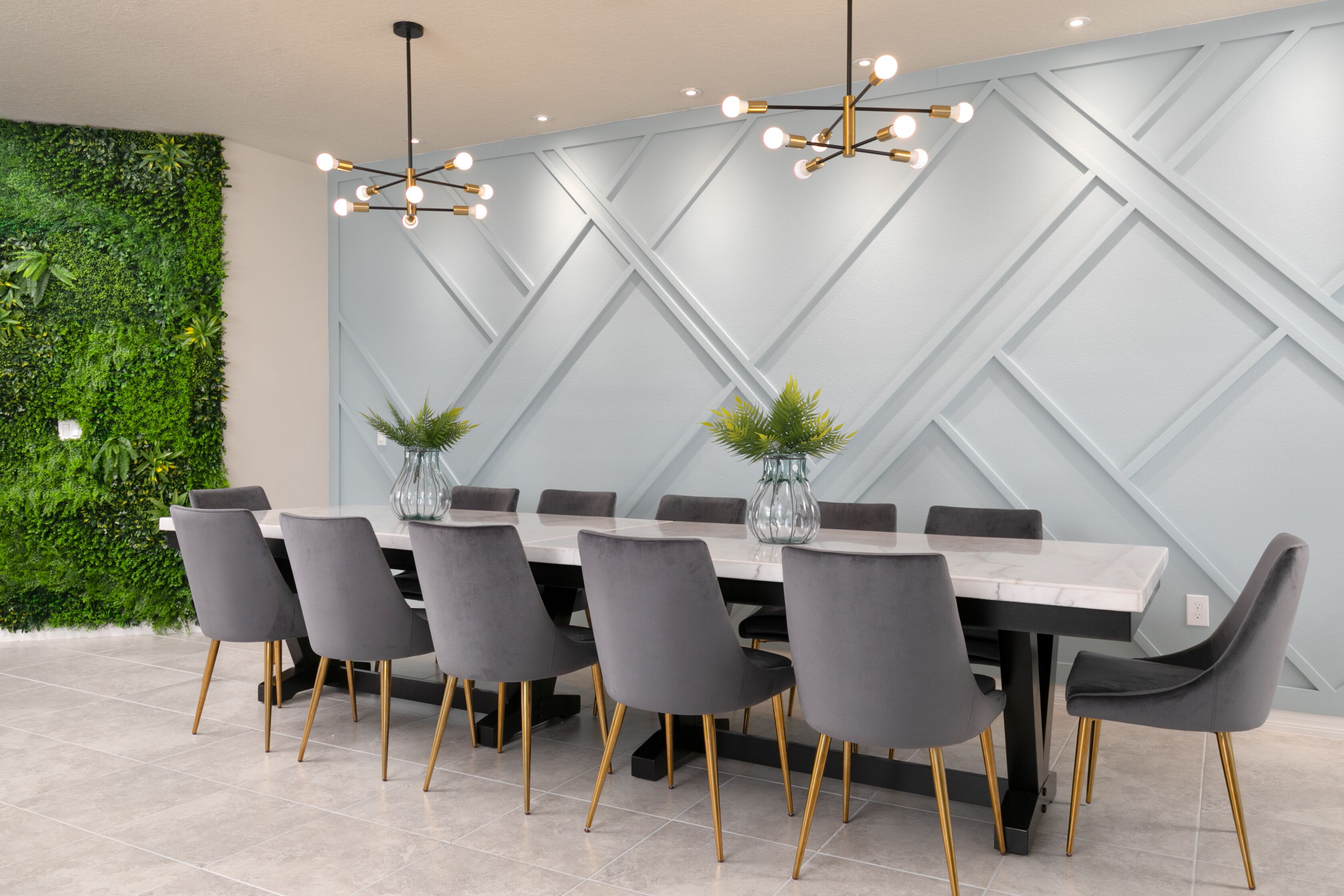 A modern dining area with a large rectangular table, six grey upholstered chairs with gold legs, and two pendant lights with multiple bulbs.