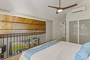 A bedroom with a bed, a ceiling fan, a balcony with a railing, and a view of the outdoors.