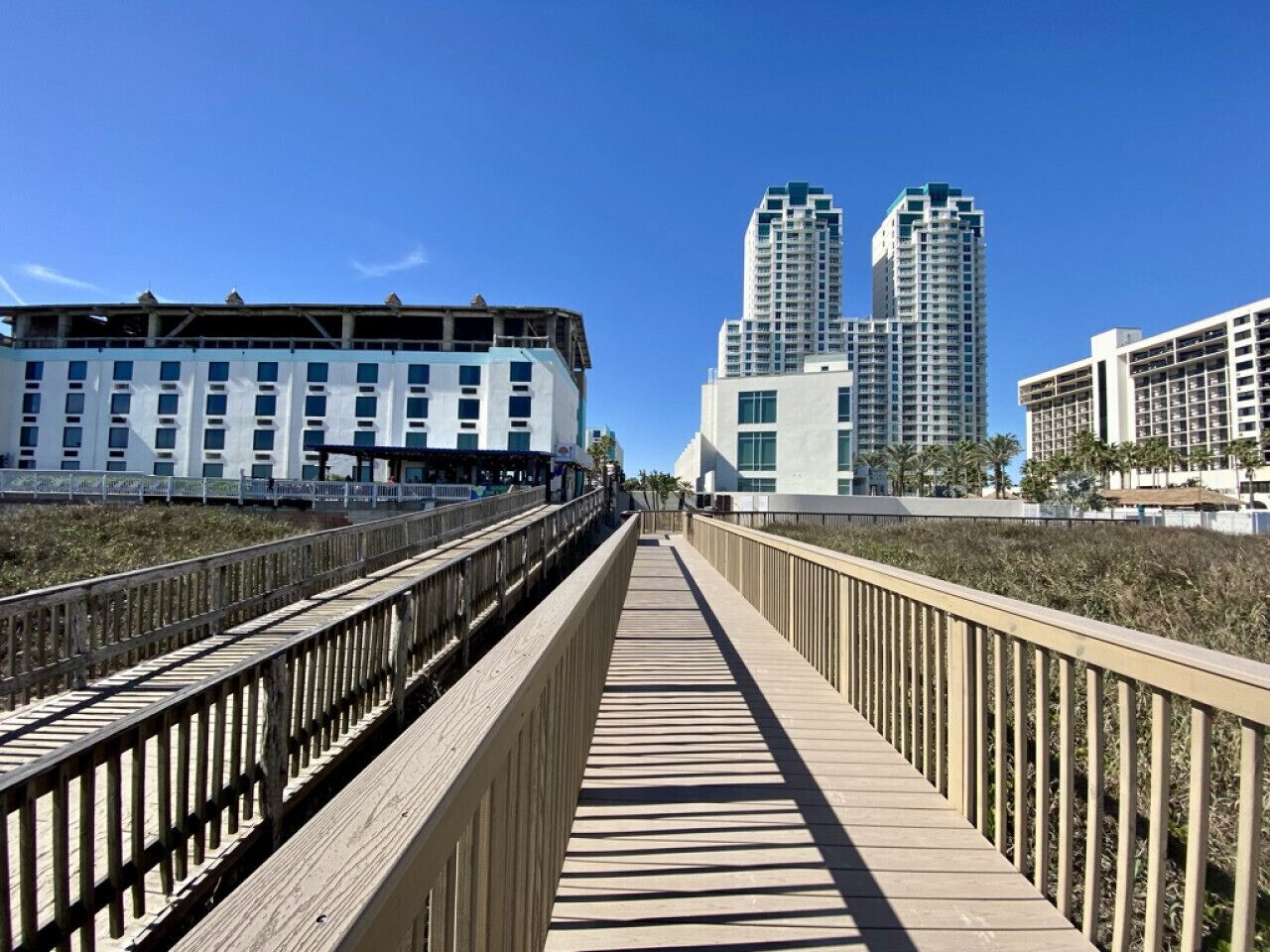 Un paseo marítimo de madera que conduce a un hotel frente a la playa, con arquitectura moderna y edificios altos al fondo.
