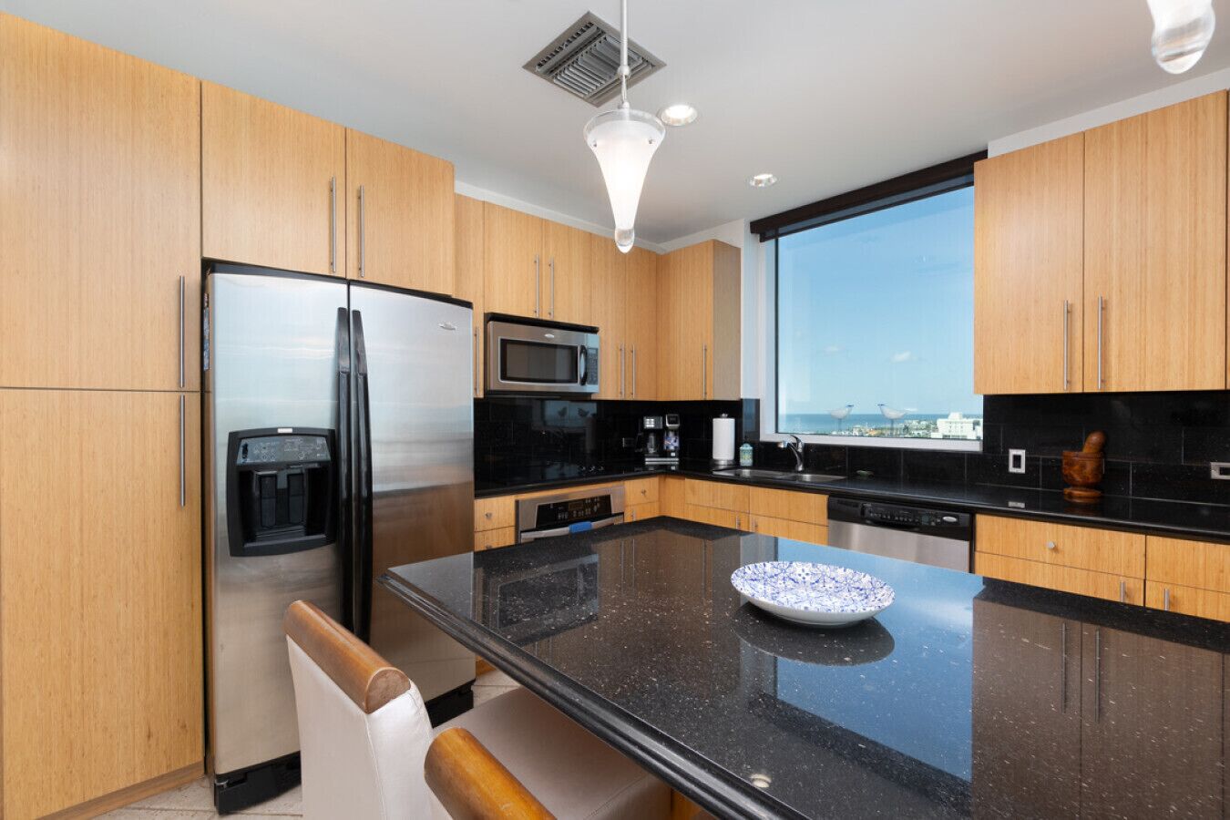 A modern kitchen with wooden cabinets, a stainless steel refrigerator, a microwave, and a large window offering a view.