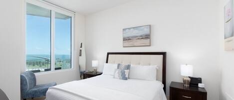 A hotel room with a large bed, a view of the ocean, a ceiling fan, and a nightstand with a lamp.