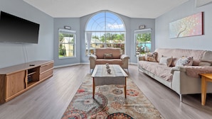 A spacious living room with a large window, a ceiling fan, a flat-screen TV, a wooden entertainment unit, and a patterned rug.