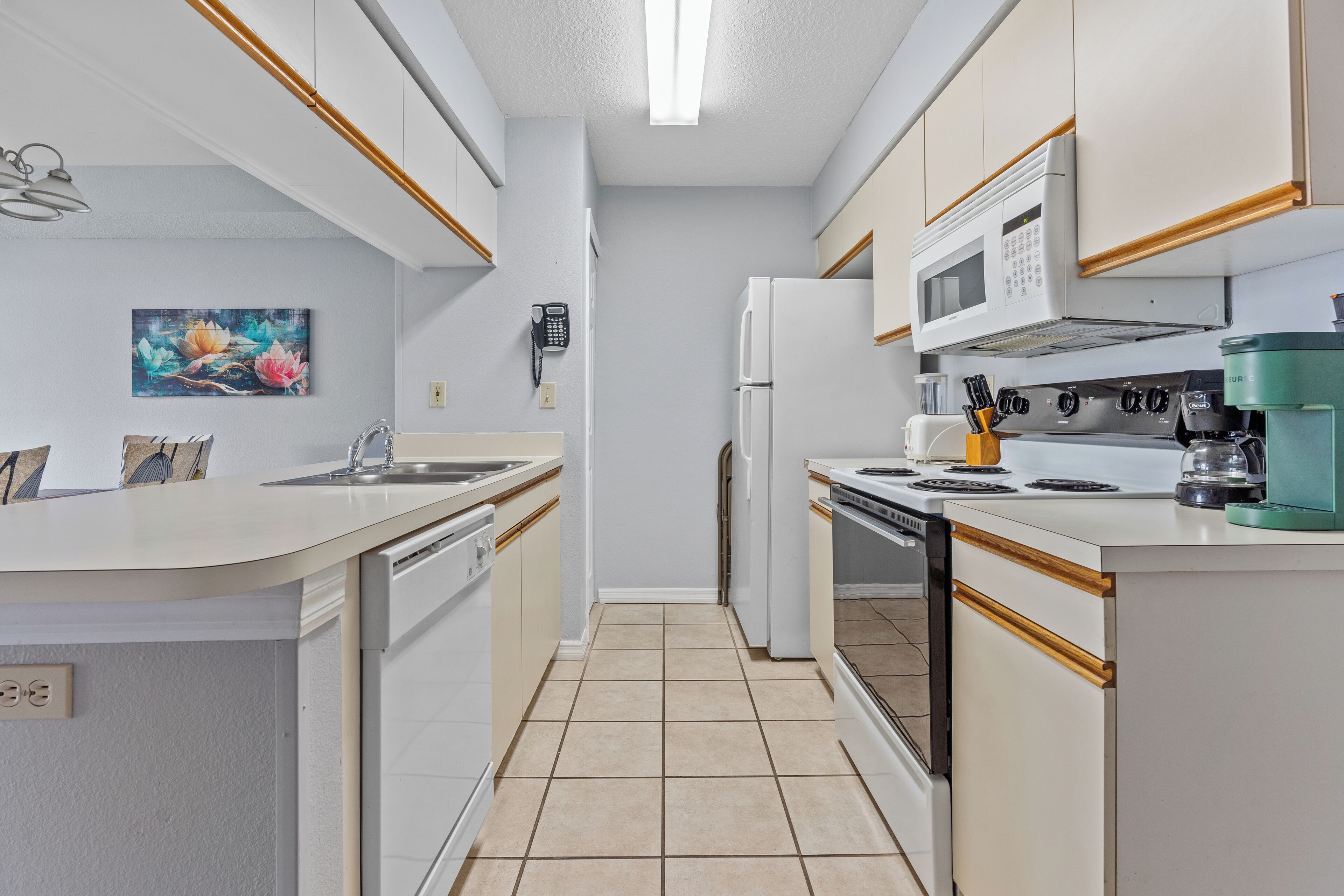 A modern kitchen with white cabinets, a built-in oven, microwave, and coffee maker. There is a sink and a painting on the wall.