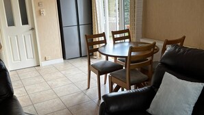 Ein Wohnzimmer mit einem schwarzen Ledersofa, einem Esstisch mit vier Stühlen, einem schwarzen Kühlschrank und einem Fenster mit Vorhängen.
